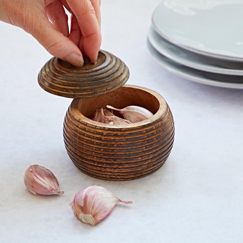 K&ouml;nnte beinhalten: Ein Knoblauchbeh&auml;lter aus Holz mit Deckel, der ein gerilltes Design aufweist. Der Beh&auml;lter ist mit Knoblauchzehen gef&uuml;llt, und daneben liegen drei Zehen auf dem Tisch.