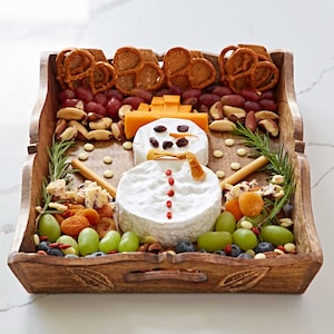 Puede incluir: Una bandeja de servir de madera con un muñeco de nieve hecho de queso y fruta. El muñeco de nieve tiene una cabeza de queso blanco, un cuerpo de queso blanco y una nariz de zanahoria. El muñeco de nieve está rodeado de pretzels, uvas, arándanos y nueces.
