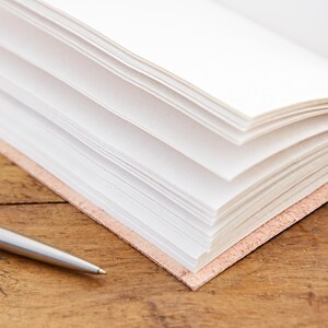 May include: A close-up of a blank, white journal with a leather cover. The journal is open and the pages are fanned out. A silver pen is lying on a wooden surface next to the journal.