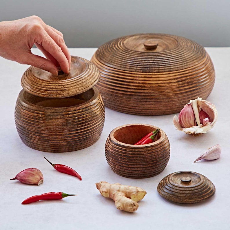 K&ouml;nnte beinhalten: Satz mit vier Holzschalen mit Deckeln, perfekt zum Aufbewahren von Gew&uuml;rzen oder anderen kleinen Gegenst&auml;nden. Die Schalen sind aus dunklem Holz mit einer gerippten Textur und haben eine nat&uuml;rliche Oberfl&auml;che. Die Deckel sind ebenfalls aus Holz und haben einen Knopf oben. Die Schalen sind auf einer wei&szlig;en Oberfl&auml;che angeordnet, mit verschiedenen Gew&uuml;rzen und Kr&auml;utern, die um sie herum verstreut sind.