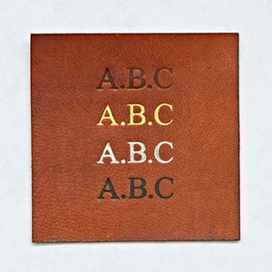 May include: A close-up of a brown leather patch with the letters "A.B.C." embossed in black, gold, silver, and black.