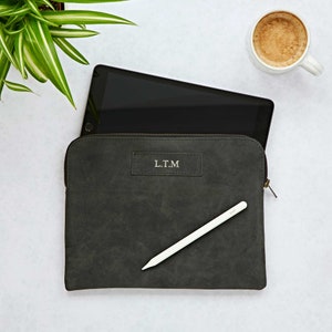 Könnte beinhalten: Eine dunkelgraue Tablettasche aus Wildleder mit Reißverschluss und einem weißen Stift. Die Tasche hat die Initialen "L.T.M." auf der Vorderseite geprägt.