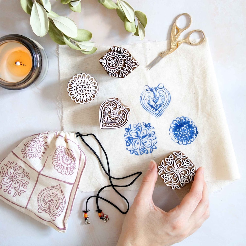 May include: A collection of wooden block stamps with intricate floral and heart designs, alongside blue ink impressions on fabric. A drawstring bag with matching patterns and a pair of gold scissors are also present. A hand is holding one of the stamps.