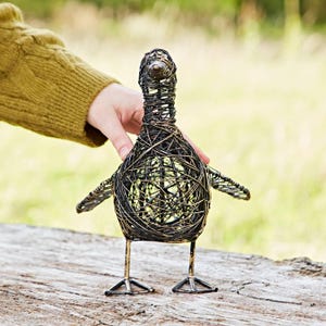 May include: A wire-wrapped metal sculpture of a duck with a black and gold finish. The duck is standing on a wooden surface.