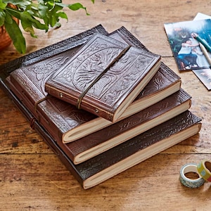 Könnte beinhalten: Ein Stapel brauner Lederjournale mit geprägten floralen und keltischen Knotenmustern. Die Journale haben einen Riemenverschluss und cremefarbene Seiten. Sie liegen auf einer Holzoberfläche.