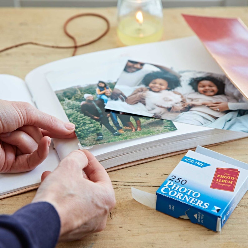 May include: A box of 250 acid-free photo corners, labeled "The Photo Album Company", with a white corner sticker partially pulled out of the box. The box is on a wooden surface with a photo album and photos.
