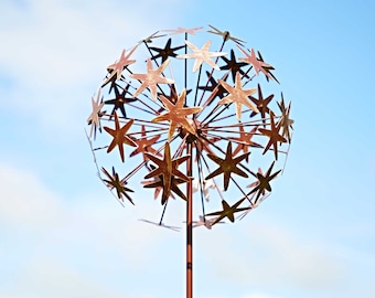 Op de afbeelding: Een metalen tuinsculptuur in de vorm van een paardenbloem met een koperen afwerking. De sculptuur heeft een ronde bol met meerdere stervormige uitsnijdingen en een lange, dunne steel.