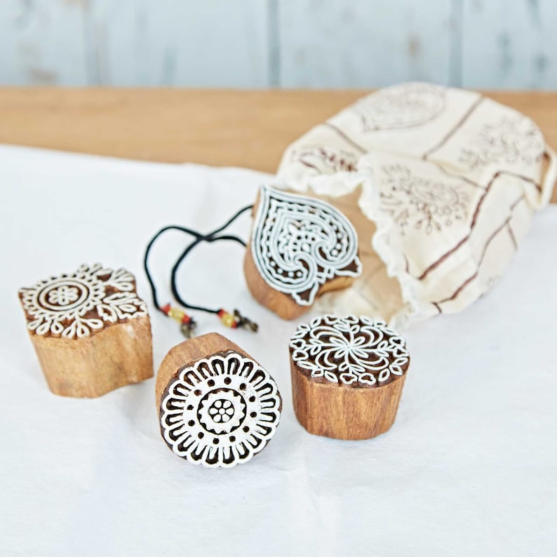 May include: Four wooden block stamps with intricate white designs, including floral and heart shapes. A drawstring bag with a matching pattern sits behind the stamps. The stamps are made of natural wood.