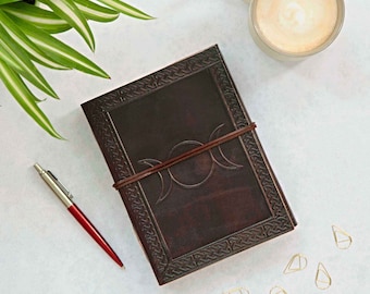 May include: A brown leather journal with a triple moon symbol embossed on the cover. The journal has a leather strap closure and is surrounded by a plant, a pen, a cup of coffee, and gold paper clips.