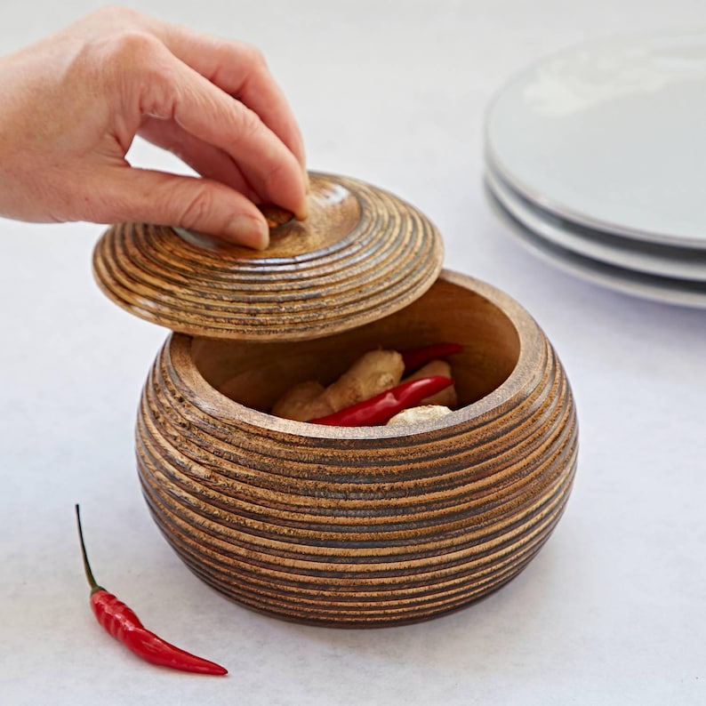 K&ouml;nnte beinhalten: Ein runder Holzbeh&auml;lter mit Deckel, der ein gerilltes Design aufweist. Der Beh&auml;lter ist mit roten Chilischoten und Ingwer gef&uuml;llt.