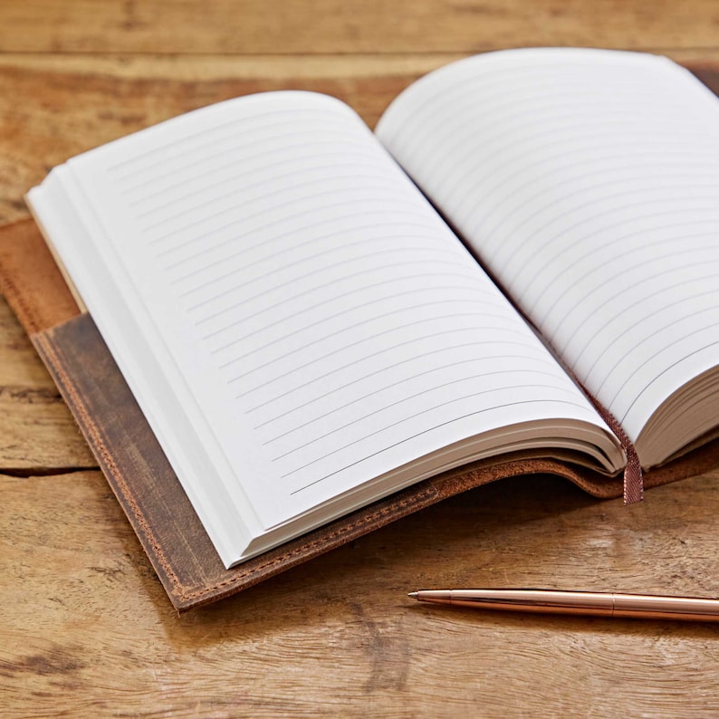 May include: A brown leather-bound journal with lined pages is open to reveal the blank pages. A gold pen is lying on a wooden surface.