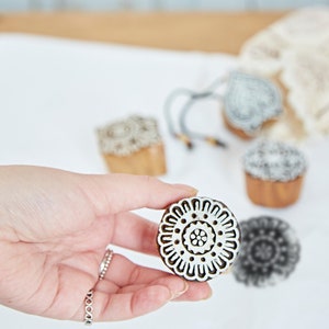 May include: A hand holding a round, wooden block with a white floral design. Other wooden blocks with various patterns are scattered on a white surface. The blocks are likely used for printing or crafting.