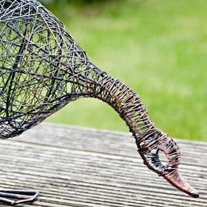 Puede incluir: Una escultura de alambre de un p&aacute;jaro con cuello largo y cuerpo redondeado. La escultura est&aacute; hecha de alambre negro y cobre y descansa sobre una superficie de madera.