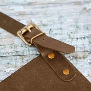 May include: Close-up of a brown leather apron with a brass buckle and two brass rivets. The apron is made of thick, durable leather and has a simple, classic design.