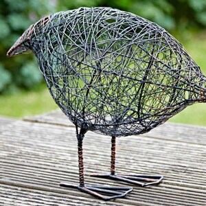 Puede incluir: Una escultura de alambre de un p&aacute;jaro con un acabado negro y cobre. El p&aacute;jaro est&aacute; de pie sobre una superficie de madera.
