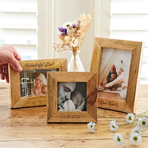May include: Three wooden photo frames with family pictures. The frames are made of light-coloured wood and have the words "Wonderful Dad" and "Our Family" engraved. One frame displays the date "07.10.2022". A small vase of dried flowers is in the background.