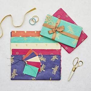 May include: A set of colorful wrapping paper with dragonfly designs. The paper is folded and stacked, with a few sheets of patterned paper on top. There are also some decorative tags and a pair of scissors.