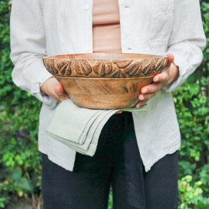 Pode incluir: Uma pessoa segurando uma tigela de salada grande de madeira com um design de folhas esculpidas. A tigela &eacute; marrom e tem um acabamento de madeira natural. A pessoa est&aacute; vestindo uma camisa branca e cal&ccedil;as pretas.