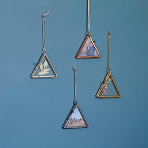 May include: Four triangular glass picture frames with silver and gold metal frames hanging from strings on a blue wall. The frames are empty except for one with a dried flower and two with photos.