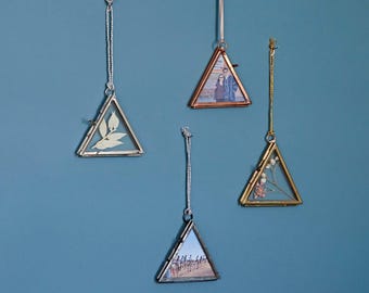 Op de afbeelding: Vier driehoekige glazen fotolijsten met zilveren en gouden metalen frames die aan touwtjes hangen aan een blauwe muur. De frames zijn leeg behalve één met een gedroogde bloem en twee met foto's.
