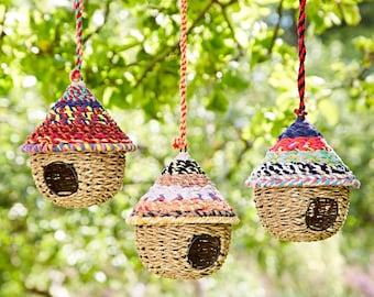 May include: Three colorful woven birdhouses hanging from a tree branch. Each birdhouse has a different pattern of colorful yarn and a woven straw base.