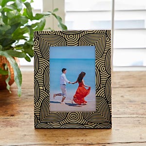 Pode incluir: Uma moldura retangular com um padrão geométrico preto e dourado. A moldura contém uma foto de um casal correndo na praia. A moldura é feita de madeira e está sobre uma superfície de madeira.