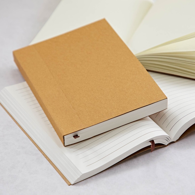 May include: A brown notebook with a lined interior is stacked on top of a white notebook with a brown cover. Both notebooks have brown ribbon bookmarks.