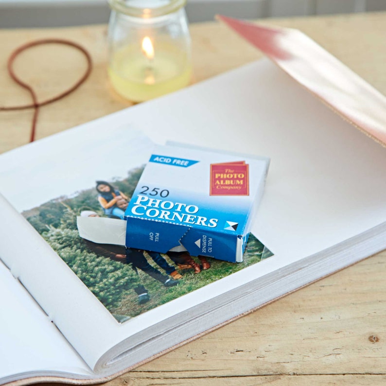 May include: A blue box of 250 acid-free photo corners, labeled "The Photo Album Company" and "250 Photo Corners". The box is open, revealing white photo corners. The box is sitting on top of a white photo album with a photo of a family in a forest.
