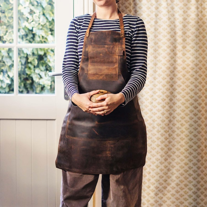 May include: A brown leather apron with a large pocket and adjustable straps. The apron is worn by a person with a striped shirt.