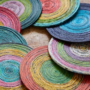 Op de afbeelding: Een verzameling kleurrijke, cirkelvormige onderzetters. Elke onderzetter is gemaakt van strak opgerolde papieren stroken in een spiraalpatroon. De onderzetters hebben verschillende kleuren, waaronder roze, groen, geel, blauw en paars. De onderzetters zijn gerangschikt op een houten oppervlak.