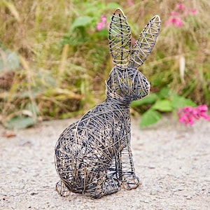 May include: A decorative wire sculpture of a rabbit, featuring a sitting pose with upright ears. The sculpture is made of interwoven black and gold-colored wire, creating a textured appearance. The rabbit is placed outdoors on a gravel surface, with greenery in the background.