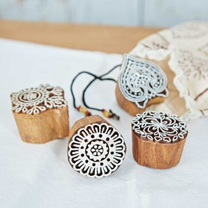 May include: Four wooden printing blocks with intricate white designs. The blocks have various shapes, including a heart and circular patterns. A black cord with beads is attached to one block. The blocks are on a white surface.