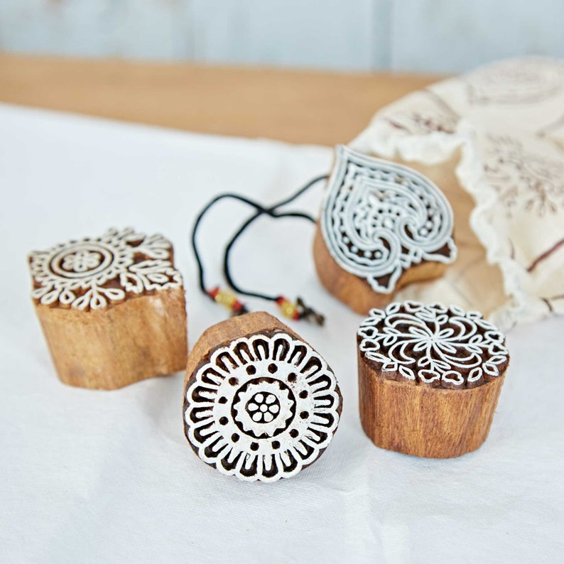 May include: Four wooden printing blocks with intricate white designs. The blocks have various shapes, including a heart and circular patterns. A black cord with beads is attached to one block. The blocks are on a white surface.