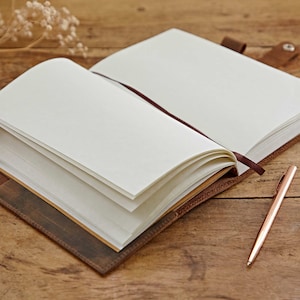 May include: A brown leather-bound journal with blank pages is open and lying on a wooden surface. A gold pen is lying next to the journal.