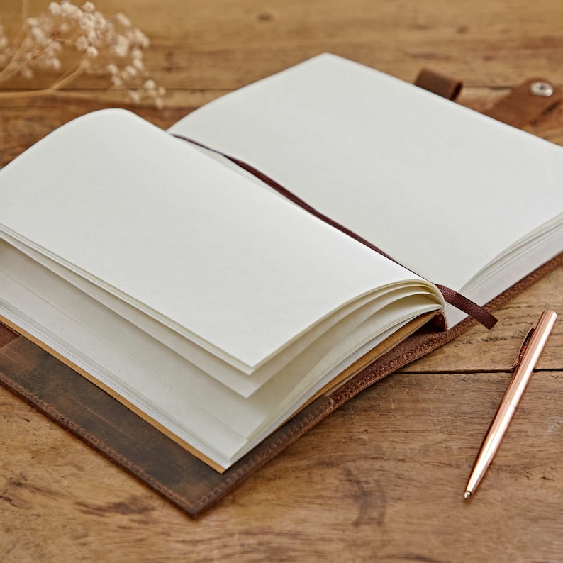 May include: A brown leather-bound journal with blank pages is open and lying on a wooden surface. A gold pen is lying next to the journal.