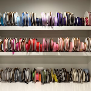 Peut inclure: Une étagère blanche à trois niveaux remplie de bobines de ruban de différentes couleurs, notamment bleu, rose, rouge et vert.