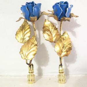 May include: Two decorative metal rose stems with blue flower heads and gold-colored leaves. The stems have a brass-colored base. The roses are set against a white background.