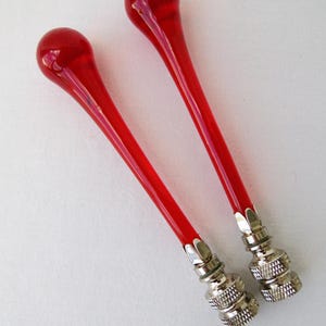 May include: Two red glass bottle stoppers with a teardrop-shaped top and a long, slender stem. The stoppers have a silver-colored metal base with a textured design. The background is a plain white surface.