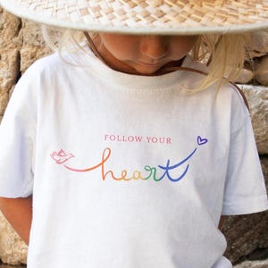 May include: A child wearing a white t-shirt with the words "Follow Your Heart" in a rainbow-coloured script. The child is also wearing a straw hat. The background is a stone wall.