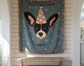 May include: A teal throw blanket featuring a dog illustration wearing a party hat. The blanket has the words "HAPPY BIRTHDAY TONTI" printed on it, with confetti shapes. The blanket has a fringed edge and is hanging on a white wall.