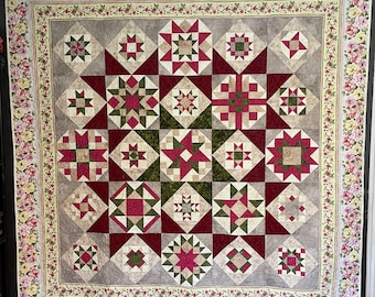 Puede incluir: Una colcha con un borde floral y un centro de patchwork. El centro está formado por 25 cuadrados, cada uno con un patrón diferente. Los patrones son principalmente geométricos y presentan tonos de rojo, verde y beige.