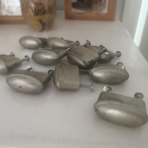 May include: A collection of ten oval-shaped cabinet knobs with a distressed silver-toned finish. Each knob features a smooth, rounded front surface and includes a metal screw attached to the back for installation, showing signs of wear.