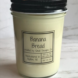 May include: A glass jar candle with a black lid and a white label that reads "Banana Bread created by Small Designs Co. 100% soy wax with cotton wick Hand-poured in Papillion, NE Net Wt. 8oz"