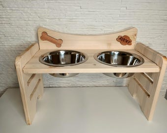 Op de afbeelding: Een houten hondenvoerstandaard met twee roestvrijstalen kommen. De standaard heeft een botuitsnijding en de naam "Rocky" is bovenaan gegraveerd.