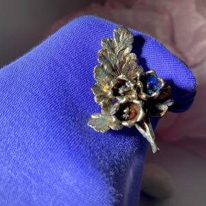 May include: A silver-toned floral brooch with detailed leaves and flower petals. The flowers have a dark center with hints of blue and orange. The brooch is set against a blue background.