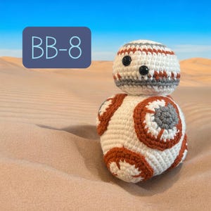 Könnte beinhalten: Eine handgefertigte BB-8-Häkel-Figur mit einem weißen Körper mit orangefarbenen und grauen kreisförmigen Details. Der Kopf ist weiß mit grauen und roten Streifen und schwarzen Knopfaugen. Der Text "BB-8" befindet sich in einem blauen Rechteck im Hintergrund.