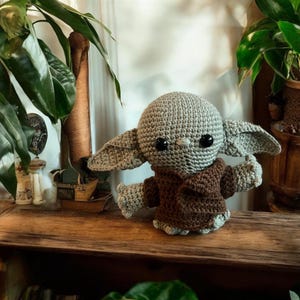 May include: A crocheted doll of a character with large ears, a gray head, and a brown robe. The doll has large black eyes and is posed with its arms outstretched. The doll is sitting on a wooden surface.