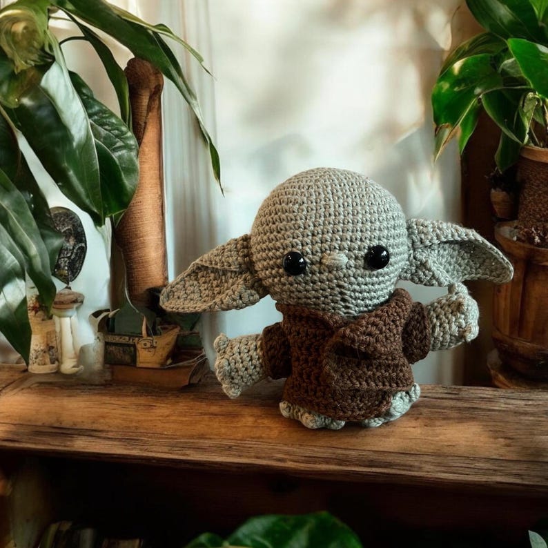 May include: A crocheted doll of a character with large ears, a gray head, and a brown robe. The doll has large black eyes and is posed with its arms outstretched. The doll is sitting on a wooden surface.