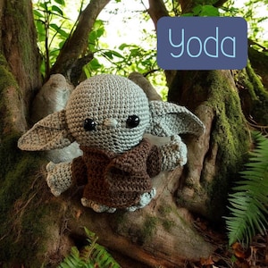 May include: A crocheted Yoda doll, featuring a gray head and large ears, wearing a brown robe. The doll has black eyes and is set against a natural background of trees and foliage. The word "Yoda" is displayed in a blue box.