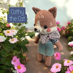 May include: A handmade crochet cat doll, Cathy, stands amidst pink flowers and greenery. The doll features a brown and white head with stitched details, rosy cheeks, and a blue dress. The text "Cathy the Cat" is in a blue box.